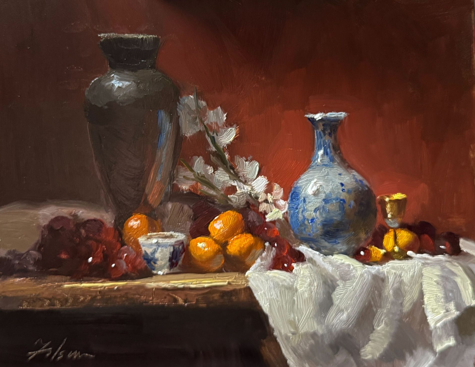 Classical still life oil painting featuring a black urn, a blue and white porcelain vase, and assorted fruit including oranges, grapes, and cherries, arranged with a gold cup and white cloth on a table against a rich red background.