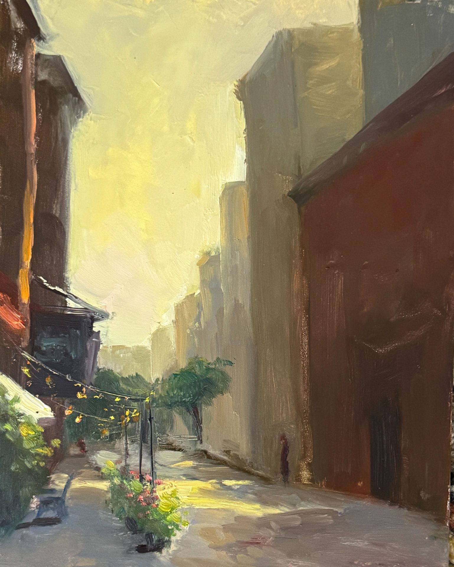 An atmospheric oil painting of a sunlit city street at golden hour, with string lights, potted flowers, and distant figures beneath towering buildings.