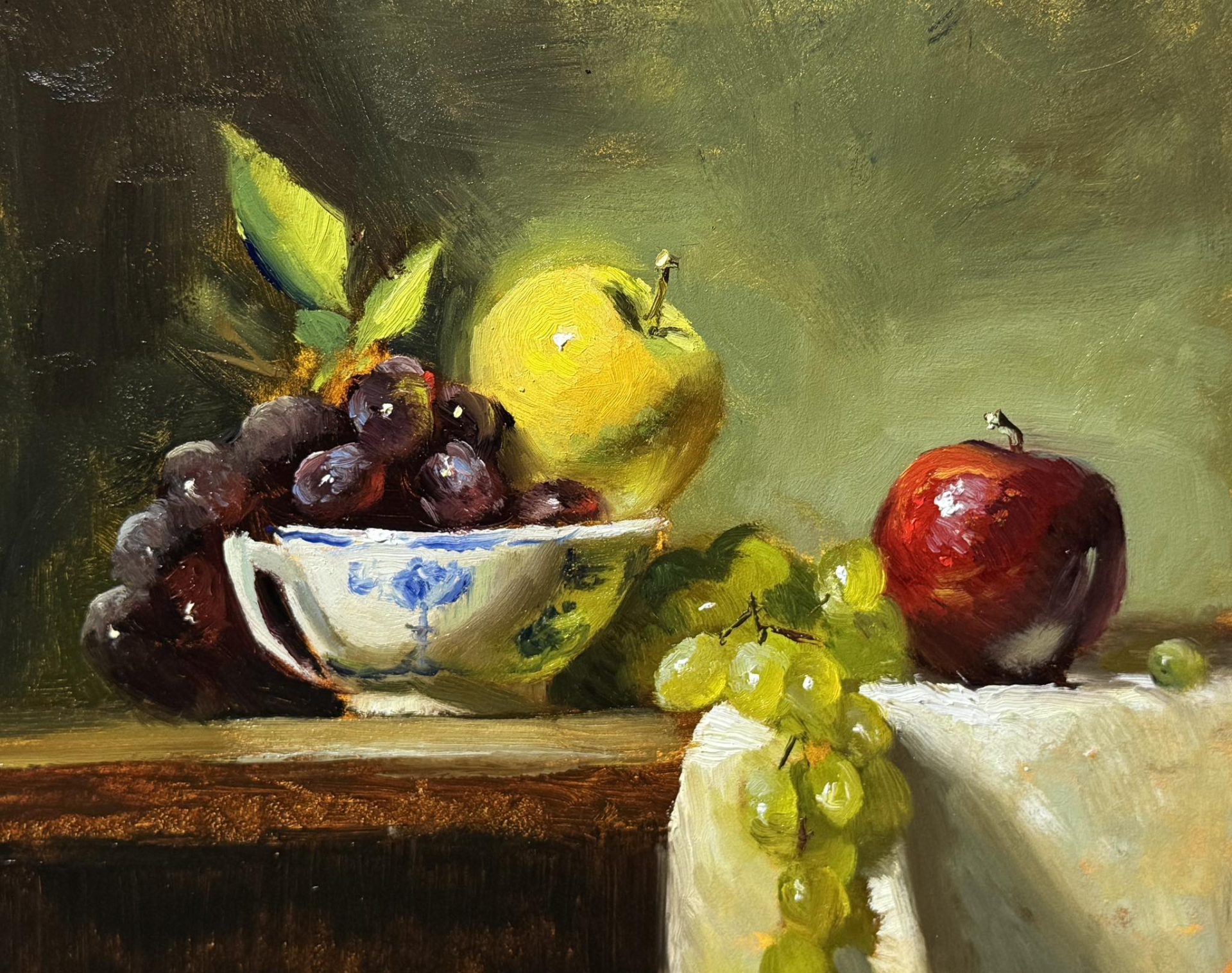Original oil still life painting of red and green apples with green grapes and dark cherries in a blue-and-white ceramic bowl, arranged on a wooden table with soft, classical lighting.